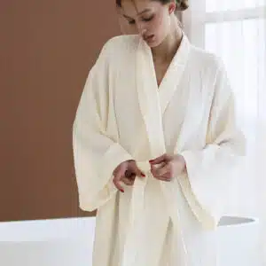 On voit une jeune femme brune dans une très belle salle de bain marron/terracotta et blanche qui porte un long peignoir léger et blanc. Elle semble distinguée et est très séduisante.