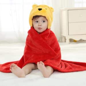 On voit un petit garçon d'environ un an et demi assis par terre dans sa chambre et emmitouflé d'un drap de bain Winnie l'ourson. La cape de bain est rouge et jaune.