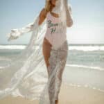 On voit une femme blonde qui déambule sur la plage avec un maillot une pièce et un peignoir en dentelle long et blanc.