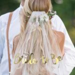 Voile EVJF bride en version doré, porté par une jeune femme blonde. On aperçoit son futur époux en arrière-plan et face à elle. Il porte une chemise blanche et une paire de bretelle marron.