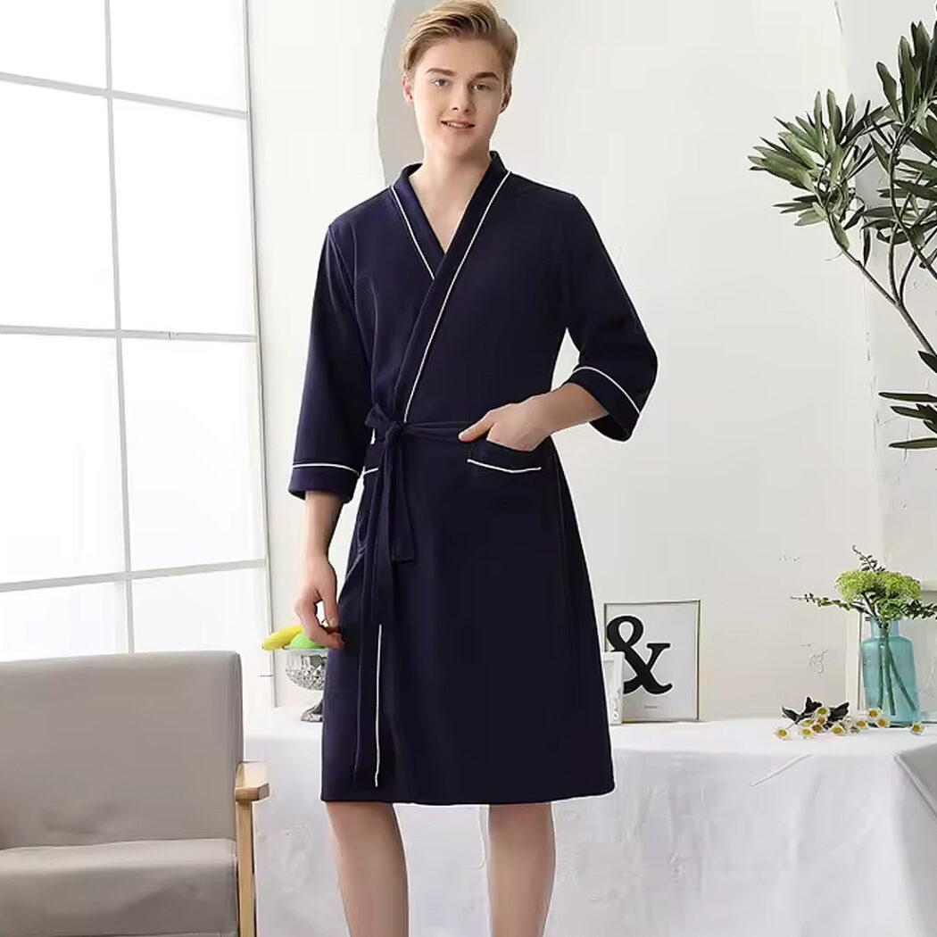 photo d'un homme portant un peignoir en nid d'abeille bleu marine mi-long avec un liseret blanc. Il se tient debout devant une chaise et une table avec une nappe blanche et a la main dans la poche.